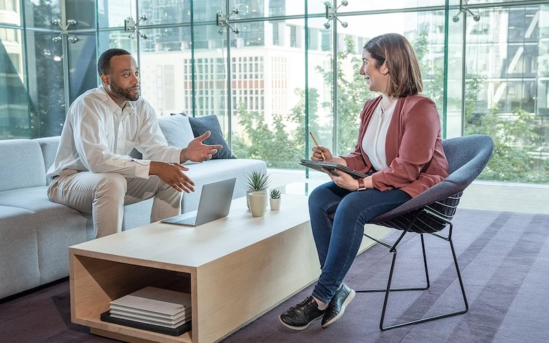 A man and a woman sit in a bright casual workspace discussing content read from a laptop on a coffee table that is between them.