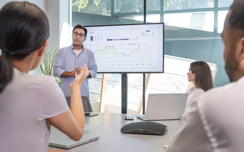 Man standing beside screen with charts presenting to a group seated around a table in a brightly lit office boardroom