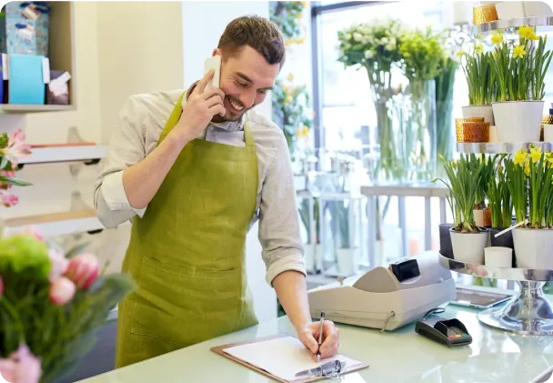 A smiling florist in a green apron talks on the phone and writes on a clipboard. The shop is filled with potted plants and flowers, with a cash register and card reader on the counter.