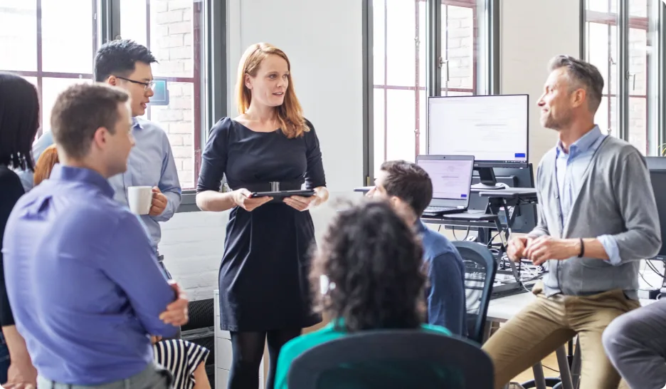 A group of colleagues meeting informally, mix of standing up and sitting down, in an office with high ceilings and large windows