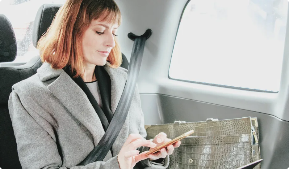 An individual sitting in a car, working from their smartphone. Three icons are visible; a clipboard, a bill and a computer monitor underneath a cloud.