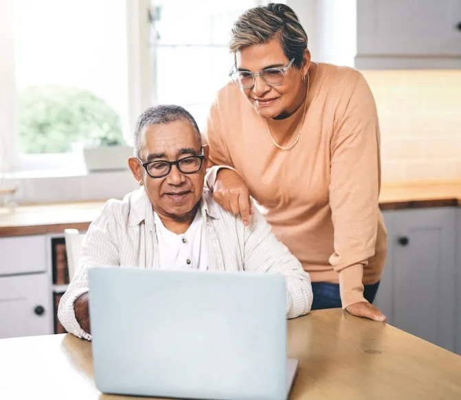 An older man seated at a kitchen able viewing a laptop screen while an older woman stands behind him