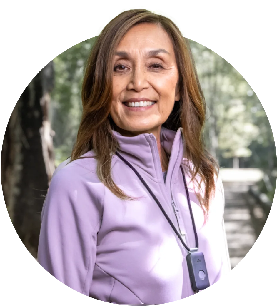 A smiling woman standing in a sunny trail wearing a a TELUS Health Medical Alert Pendant