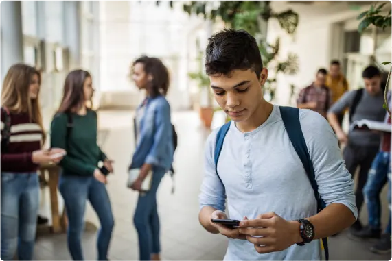 A teenager viewing a smartphone while standing in a school hallway with other teenagers in the background.