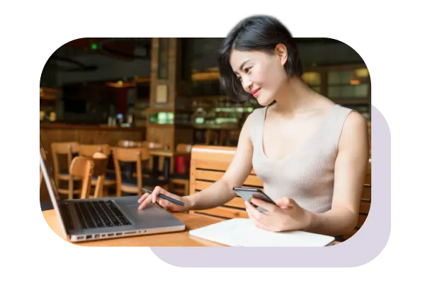 Woman working on a laptop holding a mobile phone.