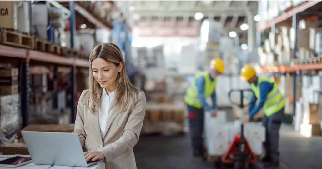 An individual viewing their laptop in a warehouse. Behind them, there are two workers in hard hats preparing to lift boxes off a pallet.