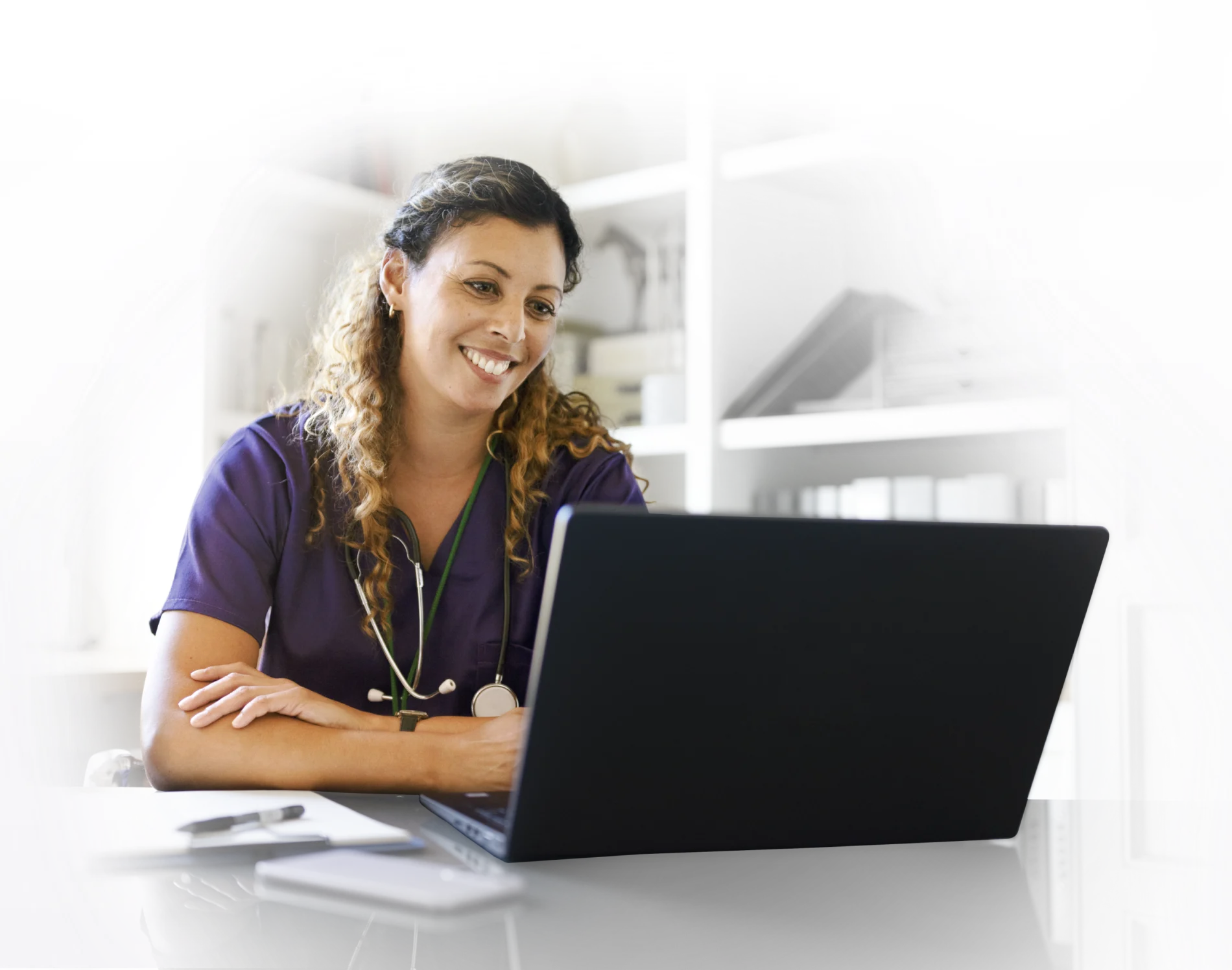 A woman seated at a desk smiling while viewing a laptop